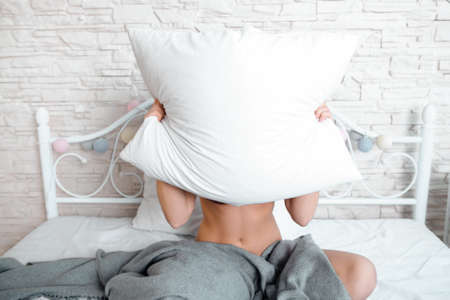 Naked woman hiding behind white pillow in bed. Sitting on bed woman covered with gray blanket and hiding her head and shoulders behind white pillow.の写真素材