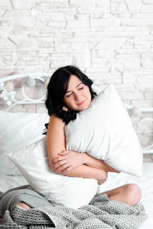 Dreaming relaxed woman hugging pillow. Beautiful woman hugging her cushion sitting on her bed in the morning. Front view on dreaming with pillow young brunette womanの写真素材