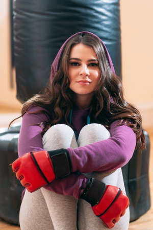 Portrait of tired woman in red boxing gloves,holding knees. Closeup photo of young female kickboxing athlete, sitting on floor near punching bag at gym, holding her kneesの写真素材