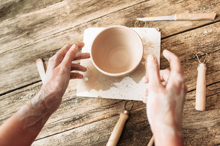 Hands above clay bowl on wooden table, artisan pov. Handmade pottery, potter working at studioの写真素材