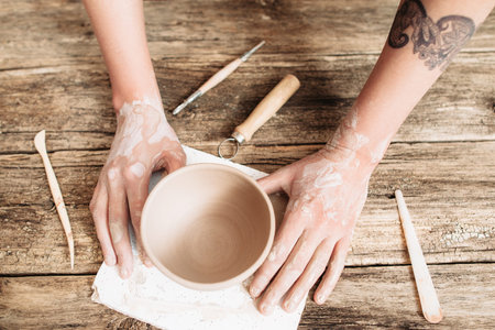 Pottery molding on wooden table top view. Artisan hands with clay bowl and special tools flat lay ,...