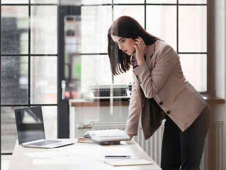Attractive business woman standing leaning on desk in office, reading her notebook.の写真素材