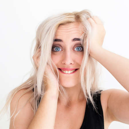 Blue-eyed blonde playing the fool at camera. Young attractive woman in black dress with widely opened eyes biting her lip and grabbing her headの写真素材