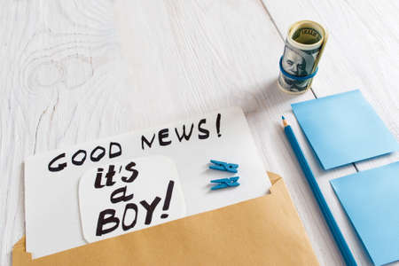 Card it's a boy with blue stickers, pencil and money, close-up. Note with congratulation about newborn child gender, white wooden background, free spaceの写真素材