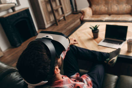 Relaxed man enjoying virtual reality glasses at home. Guy having rest in vr headset in front of laptop, back view. Modern technology, innovation, cyberspace, entertainment conceptの写真素材