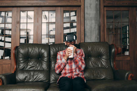 Man drinking cocktail while using vr glasses at home. Guy enjoying movie in virtual reality headset and his smoothie in ceramic glass, free space. Cinema at home, innovation, entertainment conceptの写真素材