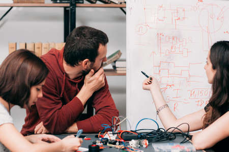Wiring diagram discussing at laboratory. Young engineers creating new model of electronic construction, experimental research of ways of connecting cables on schemeの写真素材