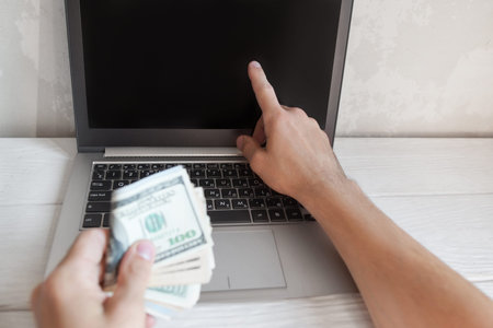 Man shows something on laptop blank screen with batch of american money in other hand. Income...