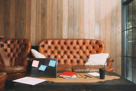 Office working place of unsuccessful worker. Wooden desk with laptop, stationery, old leather sofa...