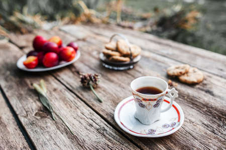Cup of coffee on wooden table outdoor, Focus on cup, blurred background of fresh plums, cookies and green nature environmentの写真素材
