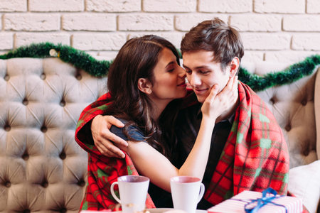 Happy couple sitting with cups of hot chocolate. Young woman kissing her man into cheek while sitting in cafe at december holidays and covering with tartan blanketの写真素材