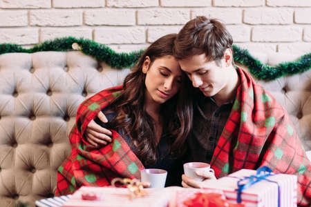 Young couple dreaming together with hot chocolate. Young man and woman hugging together covered with tartan blanket. Love, coziness, happiness, calm, gift, holidays conceptの写真素材