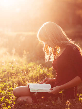 Beautiful woman reading book in nature, sun flare effect. Young attractive barefoot student in red dress studying near river at sunset. Relax, rest, education conceptの写真素材