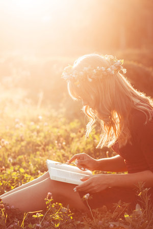Profile of girl in wreath reading book, sun flare effect. Free space on green meadow, attractive woman relaxing in nature during sunset. Rest, education, unity with nature conceptの写真素材