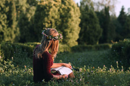 Attractive woman in wreath reading at nature, back view, copy space. Young blonde sitting with book in blooming meadow. Summer, relax, freshness, loneliness conceptの写真素材
