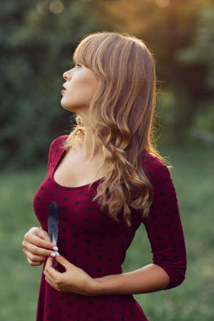 Young beautiful woman at nature. Attractive blonde in red dress holding feather and dreaming. Relax, loneliness, rest, sadness conceptの写真素材