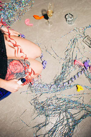 Closeup of kneeling woman with tattoos on failed new year party. Drunk girl with champagne standing on her knees on messy floorの写真素材