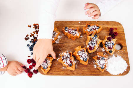 Girl put sweet cakes on board, little boy hand stealing berries in corner. Brother and sister cooking sweet treat in form of Christmas tree, top viewの写真素材