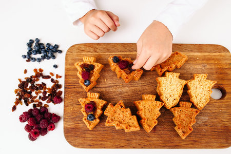 Top view on Christmas tree cakes decorating. Child hands put berries into sweet pastry on wooden board. Homemade bakery, children culinary, hobby conceptの写真素材