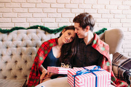 Happy couple having rest near Christmas gifts. Hugging man and woman covered with tartan blanket smiling, free spaceの写真素材
