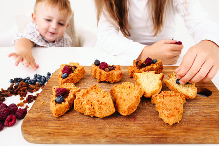 Boy stealing berries while his sister cooking. Close-up of girl cooking fruit cakes in Christmas tree form and her little brother helping herの写真素材