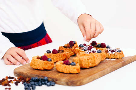 Girl decorating berry cakes with sugar powder, close-up. Young lassie cooking sweet cookies for family. Homemade bakery, children culinary, pastry making conceptの写真素材