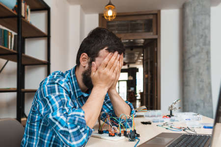Engineer closed his face in despair, free space. Side view on electrician sitting near his non working electronic construction. Failed connection, broken details, unsuccessful experimentの写真素材