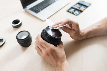 Camera lens cleaning with wet wipe, close-up. Photographer hands wiping lens on workplace . Professional photographing equipment care, technology, hobby conceptの写真素材