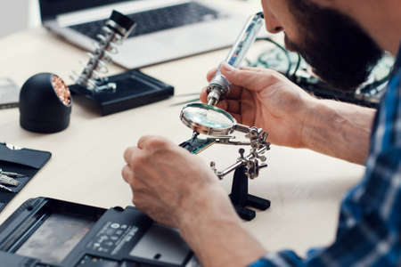 Engineer soldering microcircuit though magnifier. Electronic renovation in repair shop. Bearded repairman tinkering computer component, close-upの写真素材