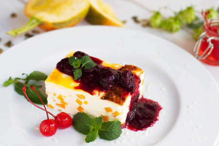Close up of cheesecake with cherry and jam on white plate. Sweet dessert serving, pumpkin and hop on blurred foreground. Autumn menu, restaurant cuisine, pastry conceptの写真素材
