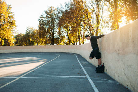 Side view on sportsman stretching arms near wall, free space. Young man doing sport exercises on open air stadium, sunset light on background, copy space on gray asphaltの写真素材