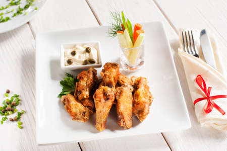 Grilled wings with vegetables and cutlery. Restaurant serving of fried chicken with creamy sauce on white plate, with fork and knife, wooden table. Junk food conceptの写真素材