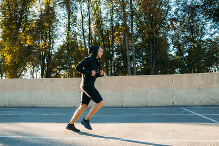 Young sportsman training on open-air stadium. Side view photo of running man in hood of sportswear, free space for text of advertisement.の写真素材