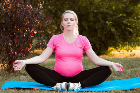 Cute pregnant woman practicing yoga outdoor. Adult expectant girl meditating at nature, sitting on karemat. Nirvana, calm, relaxation conceptの写真素材