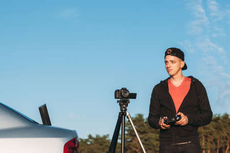 Young photographer piloting drone, free space. Attractive man moved outside by car to record portfolio with quadrocopter and camera on tripod, copy space on blue sky backgroundの写真素材