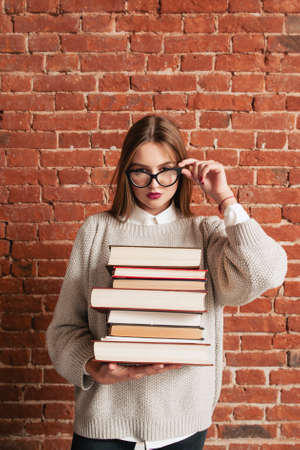 Sexy student with books looking through glasses. Attractive young woman posing with bunch of tutorials, free space on brick wall background. Challenge, sex, seduction, education, hard exam conceptの写真素材