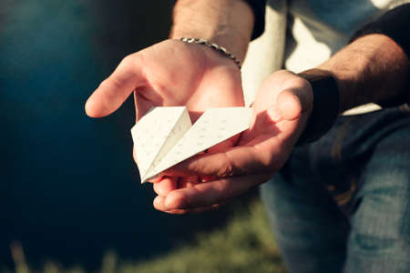 Paper airplane in male hands close-up. Man holding origami plane in palms outdoor near river. Hobby, leisure, entertainment, handmade, craft conceptの写真素材