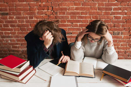 Pair of tired students preparing for exams. Young man and woman studying in library, making graduation project. Teamwork, brainstorm, education conceptの写真素材