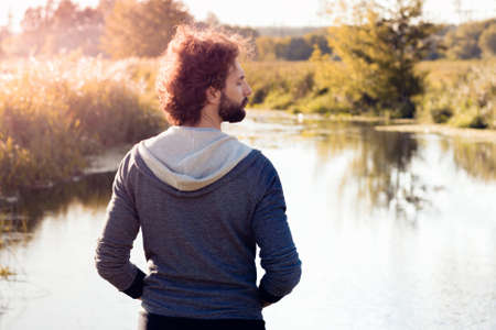 Back of man looking at river, free space. Young stylish guy enjoying beautiful nature landscape at sunrise. Relax, nature, calm, pacification conceptの写真素材