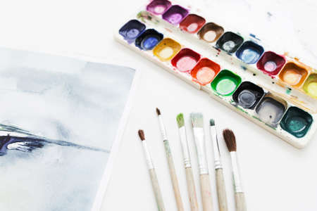 Artist workplace with drawing tools. Painted picture, set of paintbrushes and watercolor palette on white table. Art, workshop, painting, craft, creativity conceptの写真素材
