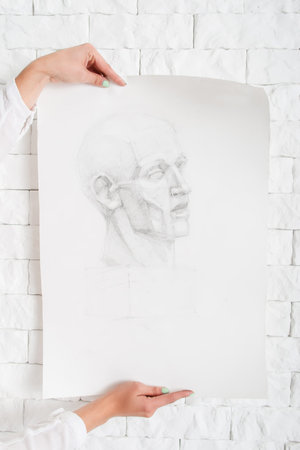Pencil portrait in artist hands against wall. Painter holding sketch of human profile near white bricks side of workshop. Art, creativity, performance, exhibition, craft conceptの写真素材
