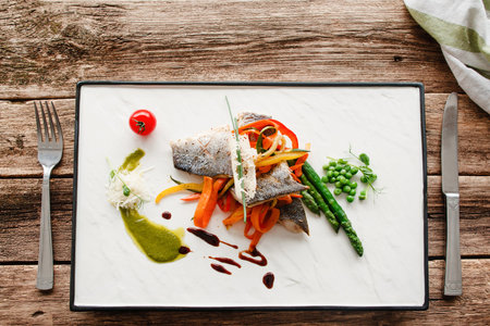 Grilled fish with fresh vegetables flat lay. Top view on dining place with roasted pangasius fillet with carrot, asparagus and pea, served with cutlery on rustic wooden tableの写真素材