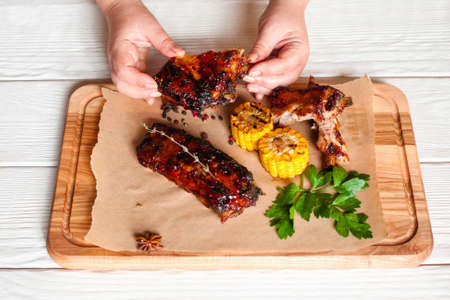 Eating grilled tasty ribs top view. Female hands holding barbecue meat above wooden board with meat and corn. Dining, enjoying picnic, junk food conceptの写真素材