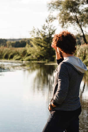 Young stylish guy enjoying calm nature landscape. Man looking at river water, back view, free space. Pacification, relax, thoughts conceptの写真素材