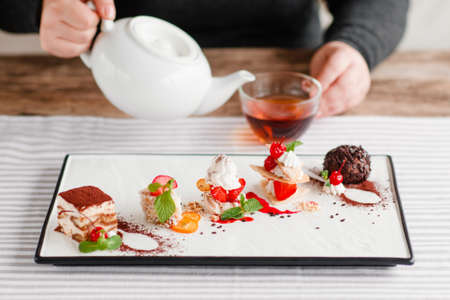Drinking afternoon tea with sweet desserts. Plate with different pieces of cakes on table, man pouring tea into cup on blurred background. Pastry, bakery, confectionery conceptの写真素材