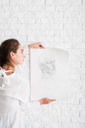 Artist hanging her pencil portrait on wall. Close-up of attractive woman decorating white brick side in her workshop with sketch. Art, creativity, hobby, talent,drawing, relax conceptの写真素材