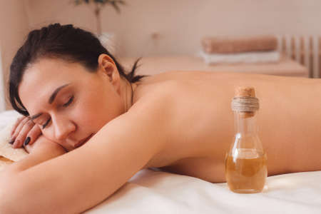 Nude woman relaxing at salon side view. Dreamy brunette having rest on massaging table with bottle of aroma oil, free space. Beauty, spa, pleasure, body conceptの写真素材