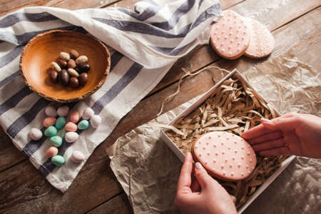 Hands put cookie into box with wooden shavings. Top view on woman preparing sweet Easter gifts, traditional treat. Homemade pastry, seasonal, holiday dessert conceptの写真素材