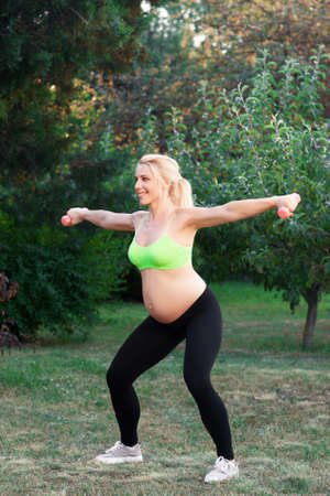 Pregnant blonde squatting with dumbbells outdoor. Sporty expectant woman exercising with sport equipment at nature. Pregnancy fitness at nature, healthy lifestyle, body care conceptの写真素材
