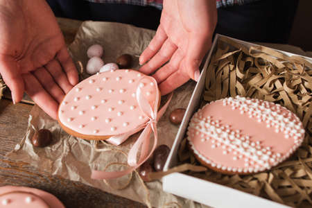 Woman packing handmade Easter cookies closeup. Top view on female hands holding sweet egg form pastry with pink glaze and gift box. Tradition, surprise, dessert conceptの写真素材